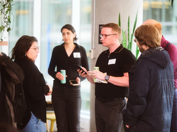 A group of people are gathered in a bright, modern indoor space, likely for a networking event or social gathering, with some individuals holding drinks and looking at their phones.