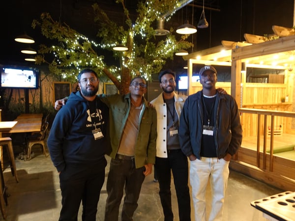A group of four men posing for a photo in an indoor social venue decorated with a lit-up tree and wooden structures.
