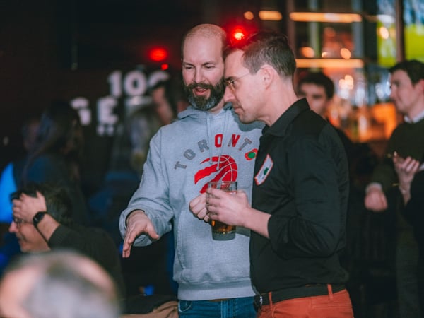 Two men are engaged in a conversation at a social gathering in a dimly lit indoor venue, with one wearing a grey Toronto Raptors hoodie and holding a drink.