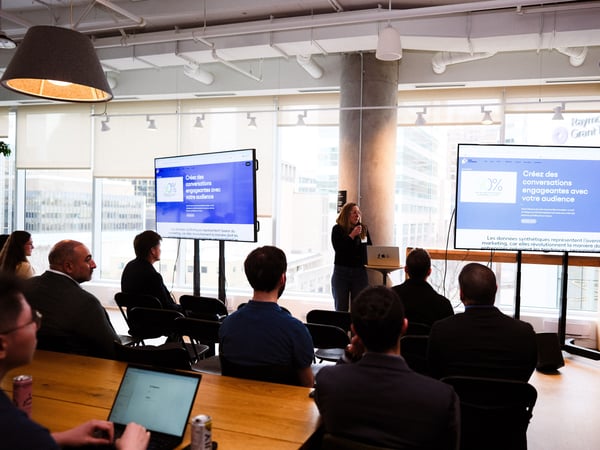 A woman stands at a podium giving a presentation to an audience in a modern office space with large windows and two digital displays showing content in French.