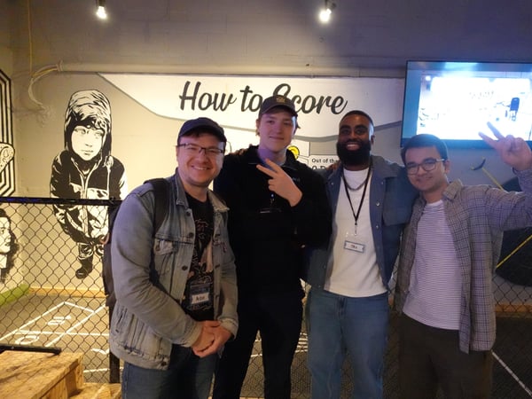A group of four young men posing for a photo in an indoor recreational space, standing in front of a wall with a mural and the text 'How to Score'.