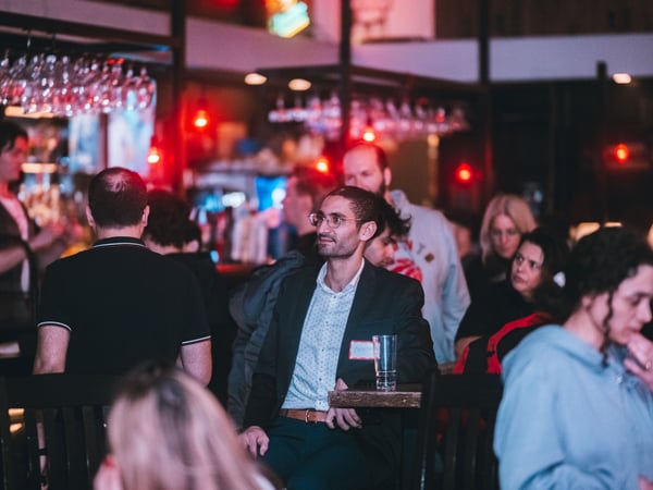 A candid photograph of a group of people at a social gathering in a dimly lit bar or lounge, featuring a man in a blazer sitting at a high table.