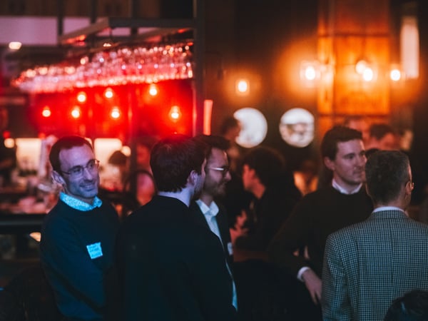 A group of people socializing in a dimly lit indoor space, likely a bar or networking event, characterized by warm red and orange ambient lighting.