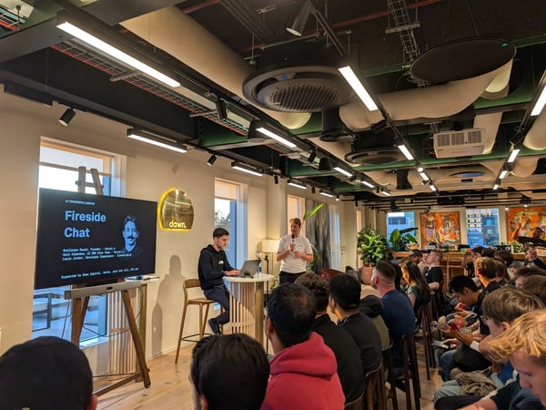 A photograph capturing an indoor 'Fireside Chat' event, likely hosted by AI Tinkerers London, featuring a speaker addressing a large seated audience in a modern office or event space.