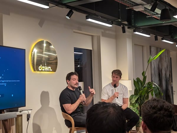 A candid photograph captures two men seated on stage holding microphones during a panel discussion or Q&A session in a modern indoor venue.