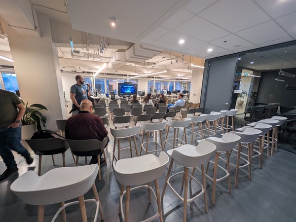 A photograph showing an indoor presentation or meeting setup in a modern office space, featuring rows of white and gray high chairs facing a large screen in the background.