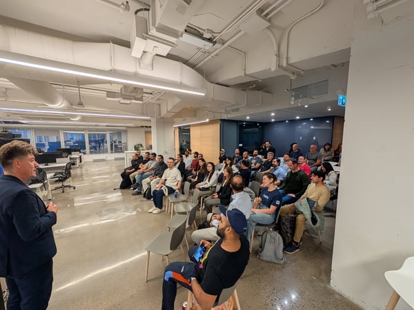A man in a dark suit is giving a presentation or talk to a large audience seated in rows of chairs within a modern, industrial-style office space.