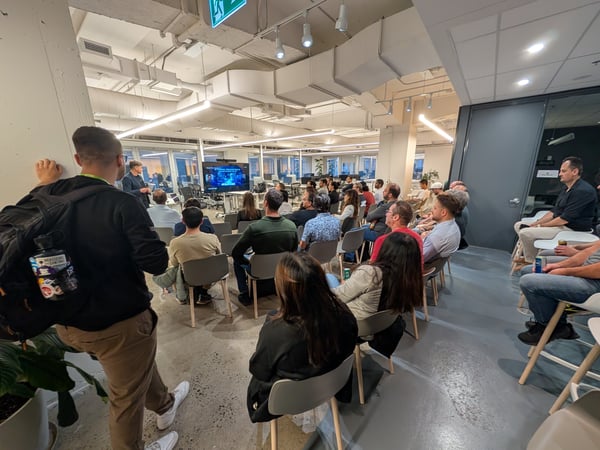 A photograph capturing an indoor event or seminar where a large audience is seated in rows listening to a speaker standing near a large monitor displaying content, set in a modern office space with exposed ceilings.