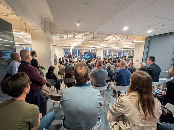 A photograph capturing a large audience attending a presentation or technical talk in a modern, open-plan office space, viewed from the back of the room.