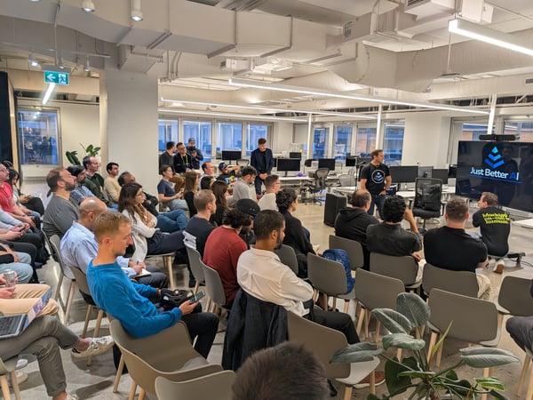 A photograph capturing a business or technology presentation taking place in a modern, open-plan office space, showing a large audience seated and facing a speaker and a screen displaying the text 'Just Better AI'.