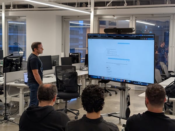 A man stands presenting content displayed on a large monitor to three audience members seated in the foreground of a modern, open-plan office space.
