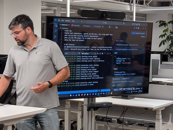 A man wearing a gray Microsoft Azure polo shirt and glasses is giving a technical presentation or demo, standing next to a large monitor displaying code, terminal output, and an AI agent chat interface in a modern office setting.