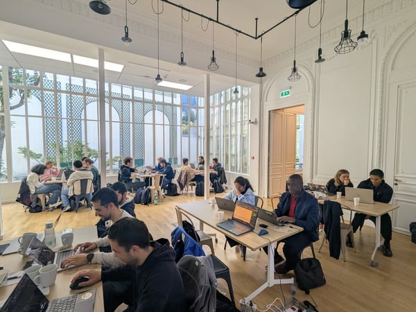 A candid photograph showing a large group of people working diligently on laptops in a brightly lit, high-ceilinged room that features ornate white architectural molding and expansive glass windows.