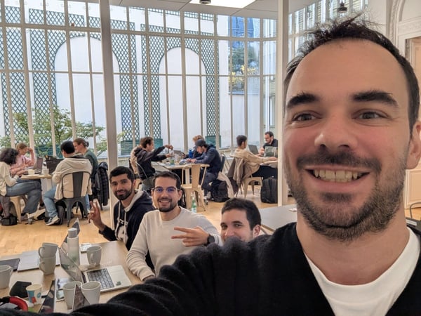 A man takes a close-up selfie in a bright, spacious co-working environment, capturing several colleagues who are either working on laptops or posing for the camera behind him.