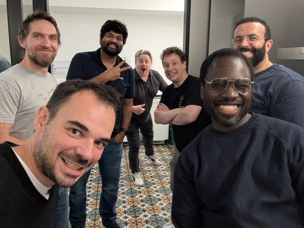 A casual group selfie featuring six men posing closely together indoors, all smiling broadly at the camera. They are standing on a floor covered with decorative patterned tiles against a plain white wall.