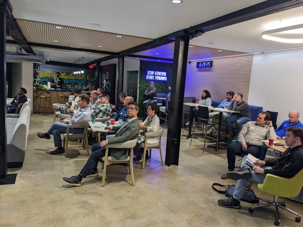 A photograph showing a group of approximately twenty people attending an indoor presentation or meeting in a modern, industrial-style venue with exposed black beams, concrete floors, and a decorative wooden slat ceiling.