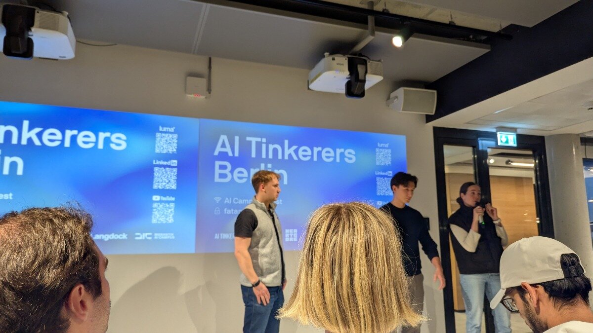 A candid photograph taken during an indoor event, showing three speakers/presenters standing in front of a large projection screen displaying the text "AI Tinkerers Berlin," while several audience members are visible in the foreground.