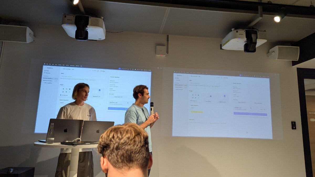 A photograph captures an indoor presentation setting where two individuals stand in front of a wall displaying dual projected screens showing software interfaces, viewed from the perspective of an audience member whose head is visible in the foreground.