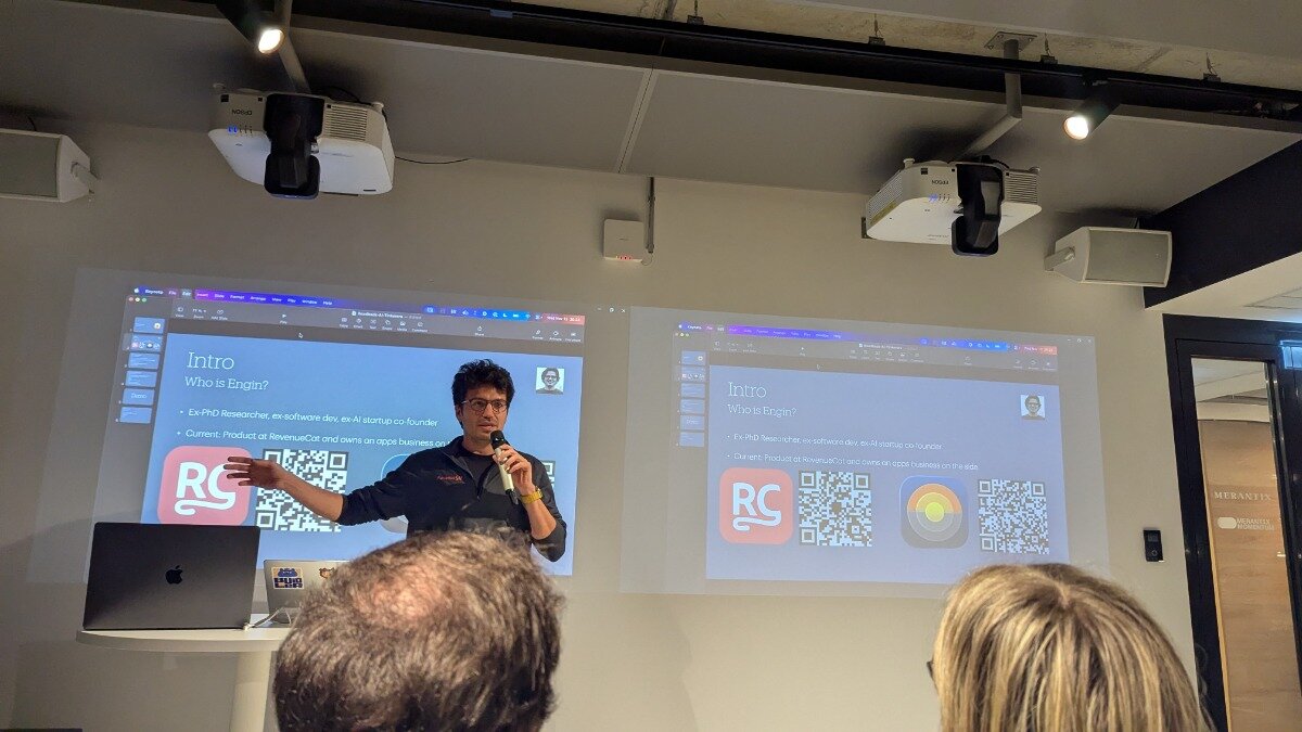 A man is giving a presentation indoors, standing between two large projected screens that display his introduction slide, while the backs of two audience members' heads are visible in the foreground.