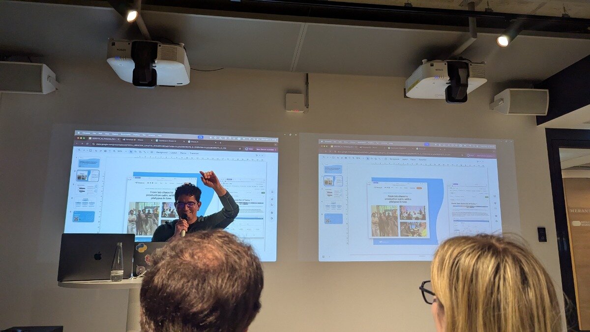 A photograph capturing an indoor presentation where a speaker is addressing an audience, with two large projection screens displaying presentation slides behind him.