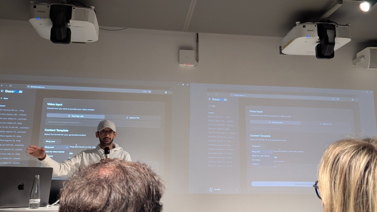 A man wearing a white hoodie and a baseball cap presents a software demonstration, standing between two large projection screens displaying a 'DocsForAI' web interface. The photo is taken from the audience's perspective, showing the backs of two attendees' heads in the foreground.