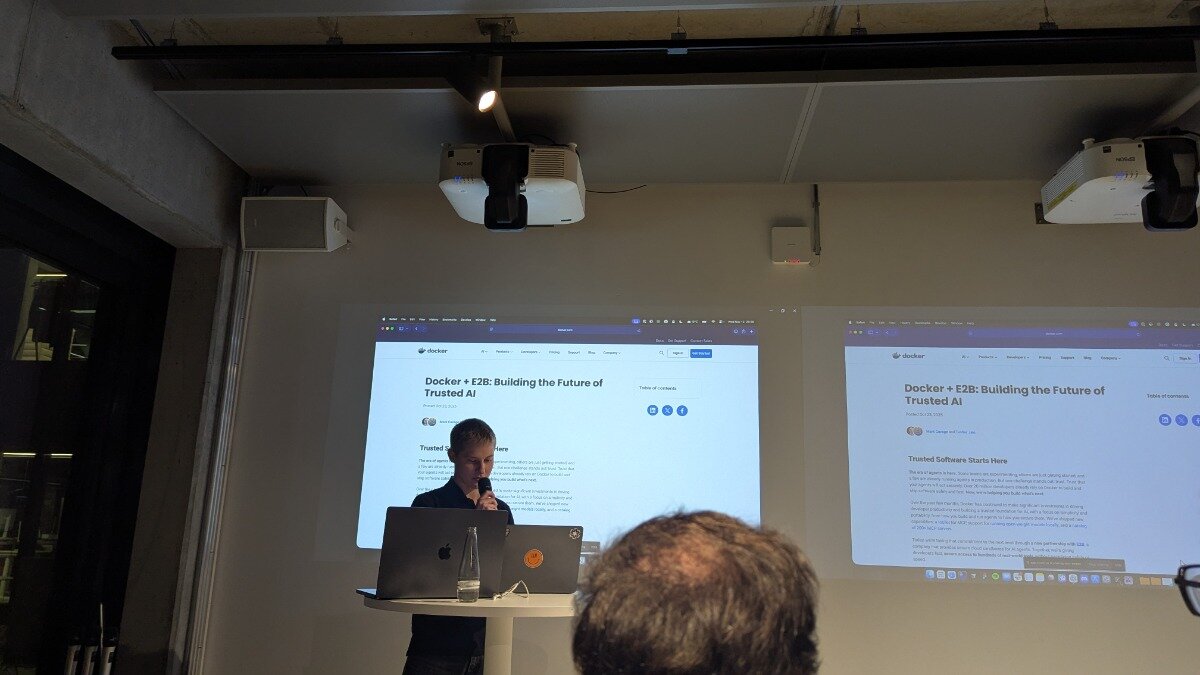 A photograph capturing a male presenter speaking into a microphone during an indoor technical event, standing in front of two large projected screens displaying content about Docker and Trusted AI.
