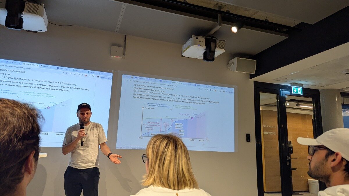 A man is giving a technical presentation indoors, speaking into a microphone while standing in front of two large projected screens displaying slides about the evolution of computing eras and entropy reduction in AI systems.