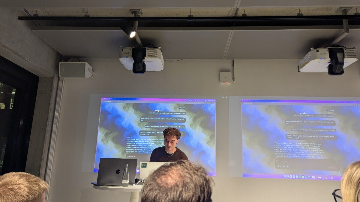 A photograph taken indoors during a presentation, showing a speaker standing in front of two large projection screens displaying chat interfaces with wavy, colorful backgrounds. The backs of the heads of two audience members are visible in the foreground.