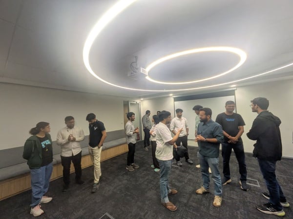 A group of approximately a dozen people are gathered in a modern office or meeting room, standing in small clusters and engaging in conversation under distinctive curved strip lighting on the ceiling.