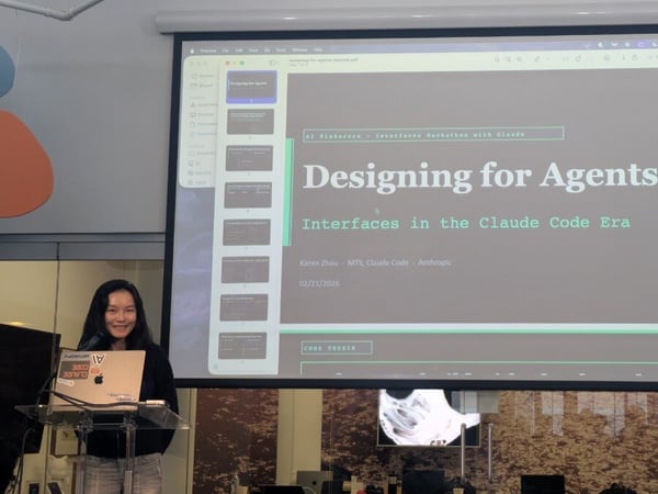 A woman stands at a transparent podium with a laptop, giving a presentation in front of a large projector screen displaying a slide titled 'Designing for Agents'.