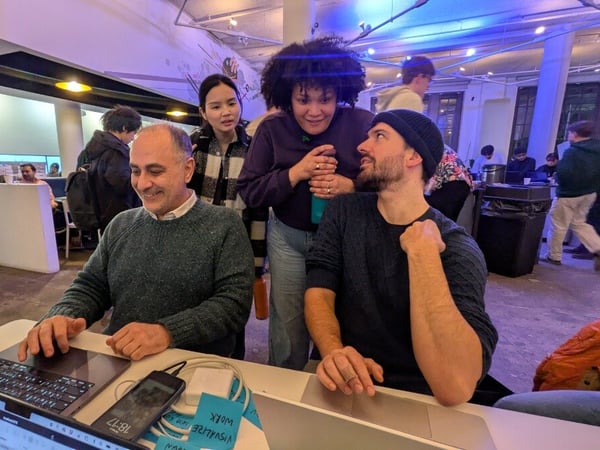 A group of people are gathered around a laptop in a modern, open-plan workspace, appearing to collaborate or debug a project together.