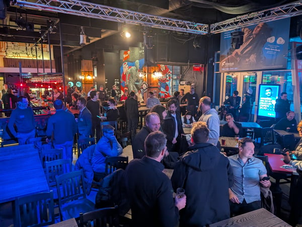 A photograph of a crowded indoor social event or bar, featuring many people standing and sitting at tables under artificial lighting.