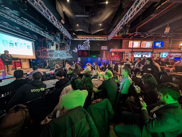 A group of people are seated in a dimly lit indoor venue, watching a presentation on a large screen while a speaker stands on a stage to the left.