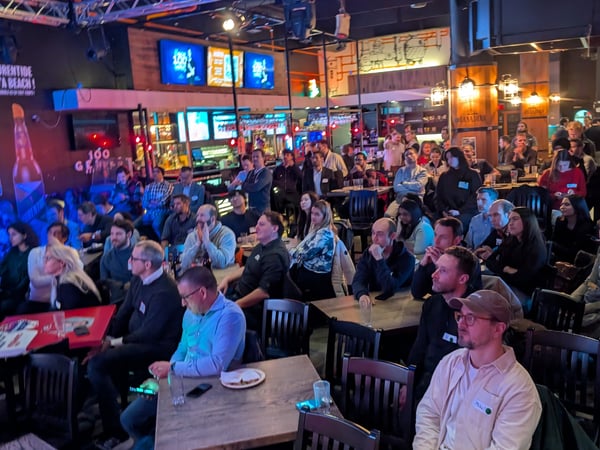 A group of people are seated at tables in a dimly lit bar or event venue, looking towards the front as if watching a presentation or performance.