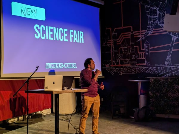 A man stands on a stage giving a presentation in front of a large projection screen displaying 'SCIENCE FAIR' and 'AI TINKERERS - MONTREAL'.