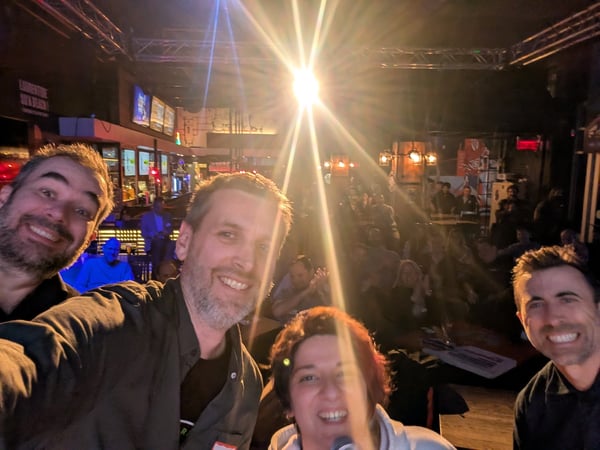 A group of four friends taking a selfie in a crowded, dimly lit bar with a prominent bright light flare in the background.