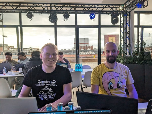 Two men are sitting at a table with laptops in an indoor venue featuring stage lighting and large windows overlooking an industrial area.