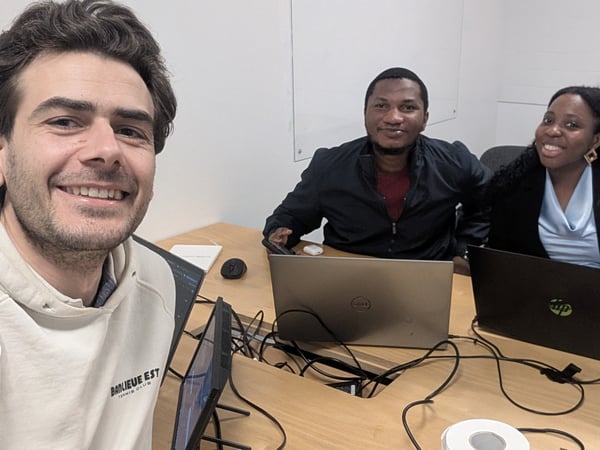 A selfie of three smiling colleagues, two men and one woman, sitting at a desk with laptops in an office setting.