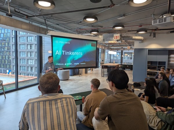A presenter stands next to a large projection screen displaying 'AI Tinkerers' and 'POLARIS' during an indoor meetup event, with an audience seated in the foreground watching.