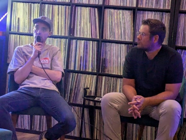 Two men are sitting on chairs in front of a massive shelf filled with vinyl records, with the man on the left speaking into a microphone.