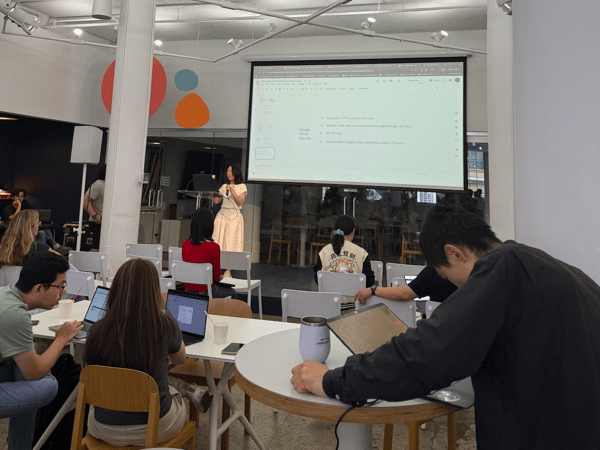 A candid photograph taken indoors showing a woman giving a presentation or workshop in a modern, brightly lit space, with the agenda projected onto a large screen behind her. Several attendees are seated at tables, many working on open laptops.
