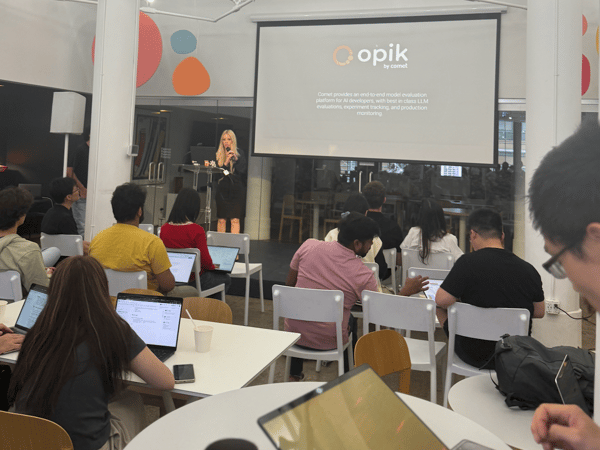 A photograph capturing an indoor presentation or lecture where a woman is speaking into a microphone on a stage while a diverse audience sits at tables, many working on open laptops. The screen behind the presenter displays the logo and descriptive text for "opik by comet."