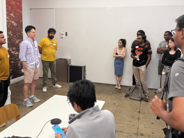 A candid photo showing a diverse group of approximately eleven people gathered indoors for a meeting or discussion, standing near a blank white wall and a patterned accent wall.