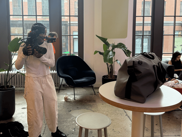 A candid photograph taken indoors showing a person operating a professional video camera, with other individuals working or standing nearby in a modern, well-lit lounge area.