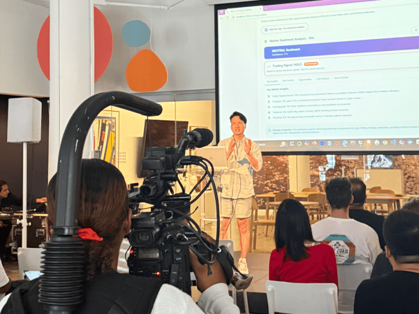 A candid photograph captured from behind a camera operator, showing a speaker presenting financial data, titled "Market Sentiment Analysis - SOL," on a large projection screen to an audience in a modern event space.
