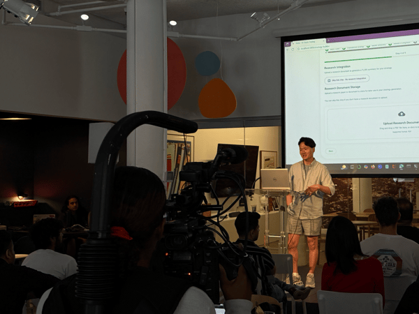 A photograph capturing an indoor technology presentation where a male speaker stands in front of a large projected screen displaying a web application interface, viewed from the perspective of the audience behind a large video camera.