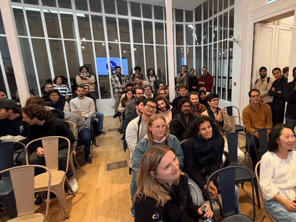 A photograph showing a large audience of people seated on metal chairs in an indoor event space, attentively watching a presentation or speaker who is out of frame to the left.