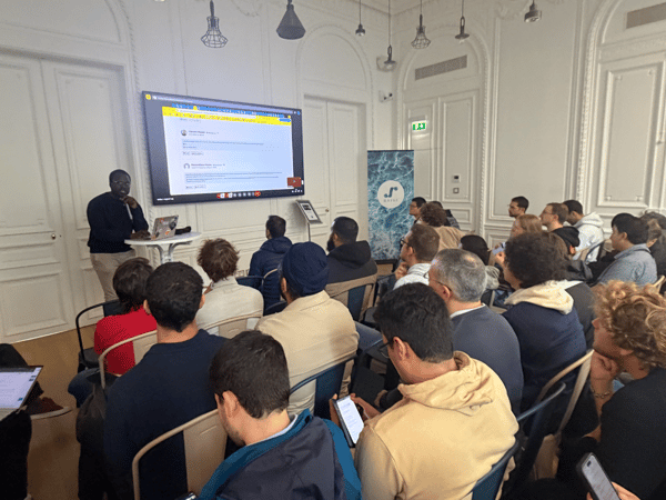 A photograph capturing a professional presentation or lecture taking place in a brightly lit, elegant room, showing a presenter standing next to a large screen while a large audience is seated, facing forward.