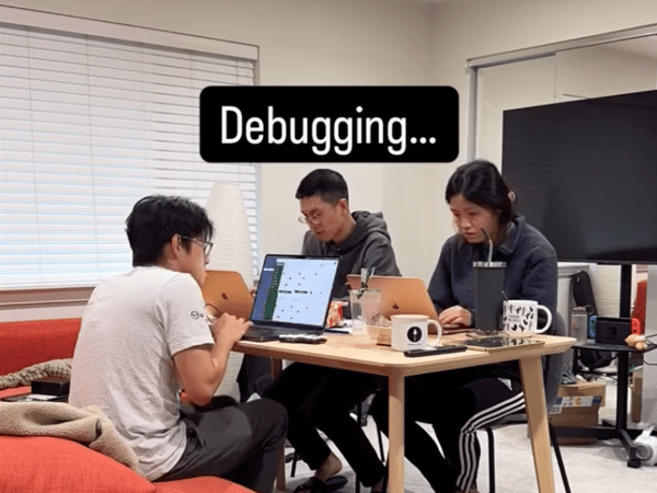 Three people are gathered around a small wooden table, intently focused on their open laptops in an indoor setting, with a text overlay indicating they are engaged in 'Debugging...'.