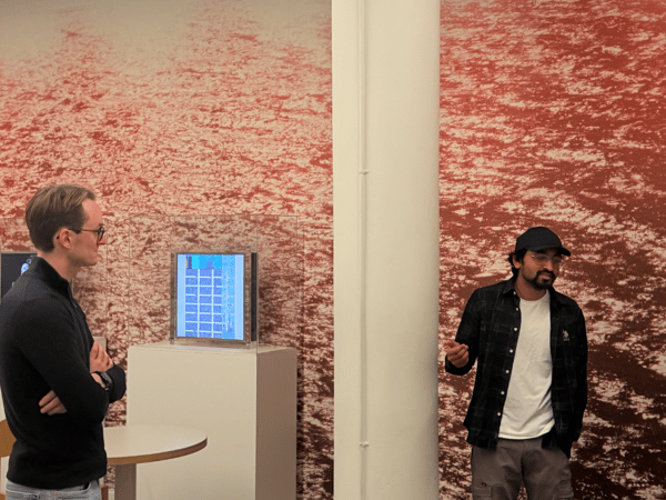 A photograph showing four men standing in an indoor space featuring a large, textured red and white wall mural, observing small digital screens displaying grid patterns mounted on white pedestals. The men appear to be engaged in a discussion or viewing an exhibition.